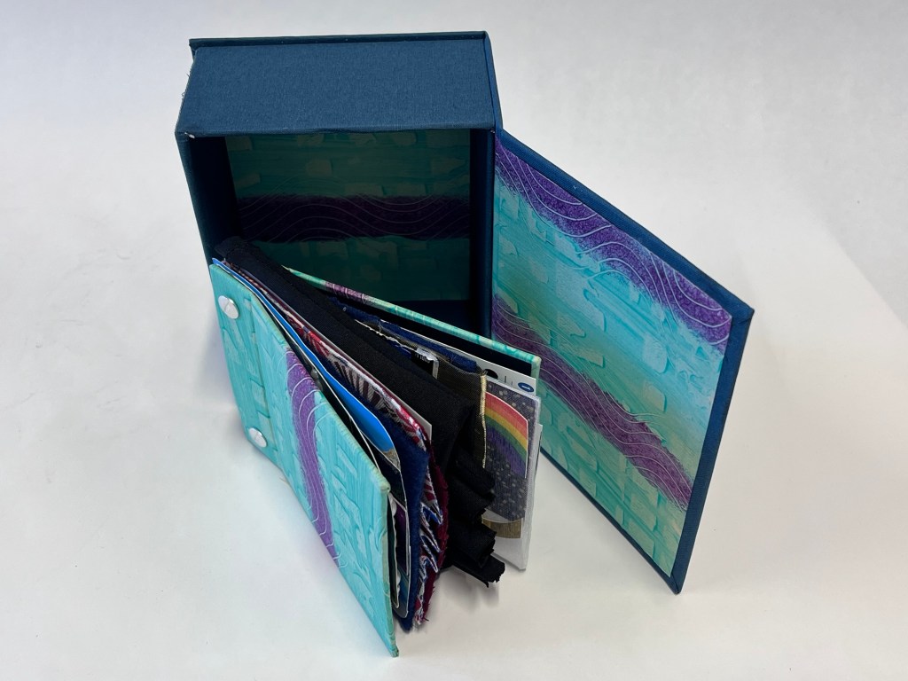 a stacked book is slightly open. it is standing outside its box. the box is blue on the outside. the inside of the box and the book cover are both striped purple and green