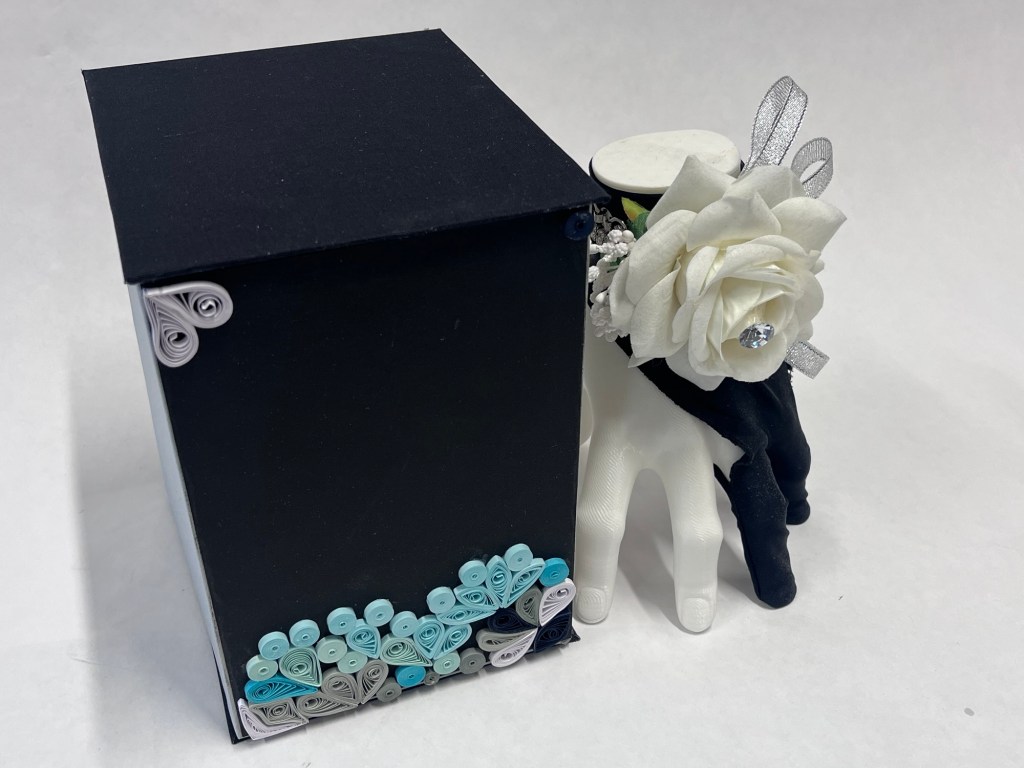 the hand sculpture, wearing a black glove and white corsage stands next to its box. the box is black and decorated in green quilled swirls